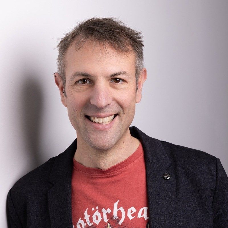 Portrait de Fabrice Sougey, souriant, en veste sombre sur un t-shirt rouge